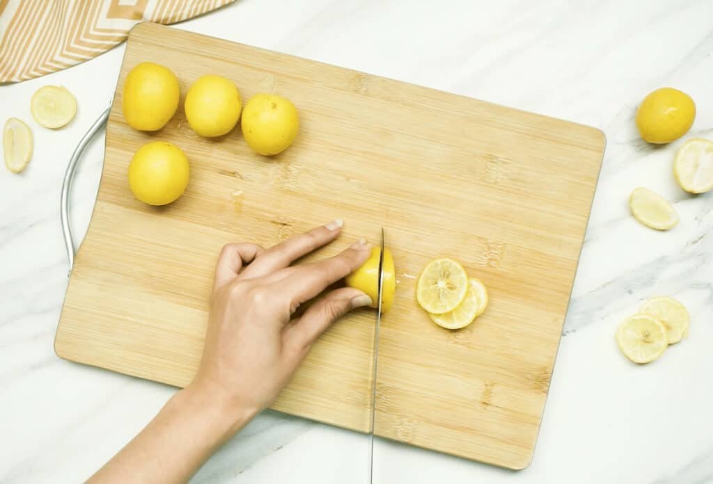 Whole lemons on a chopping board with a woman's hands slicing one lemon into thin slices