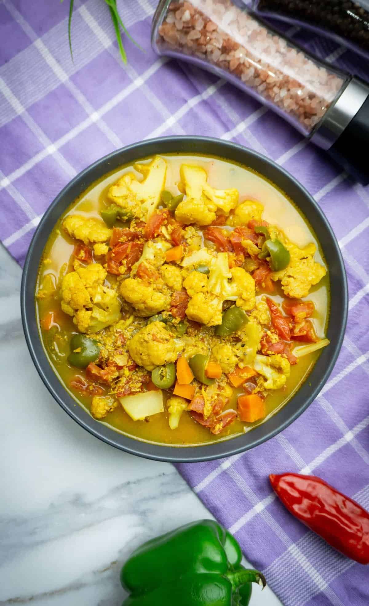 A mixed vegetable curry in a black bowl filled with cauliflower, peppers, chillies, carrots and onions in a yellow broth