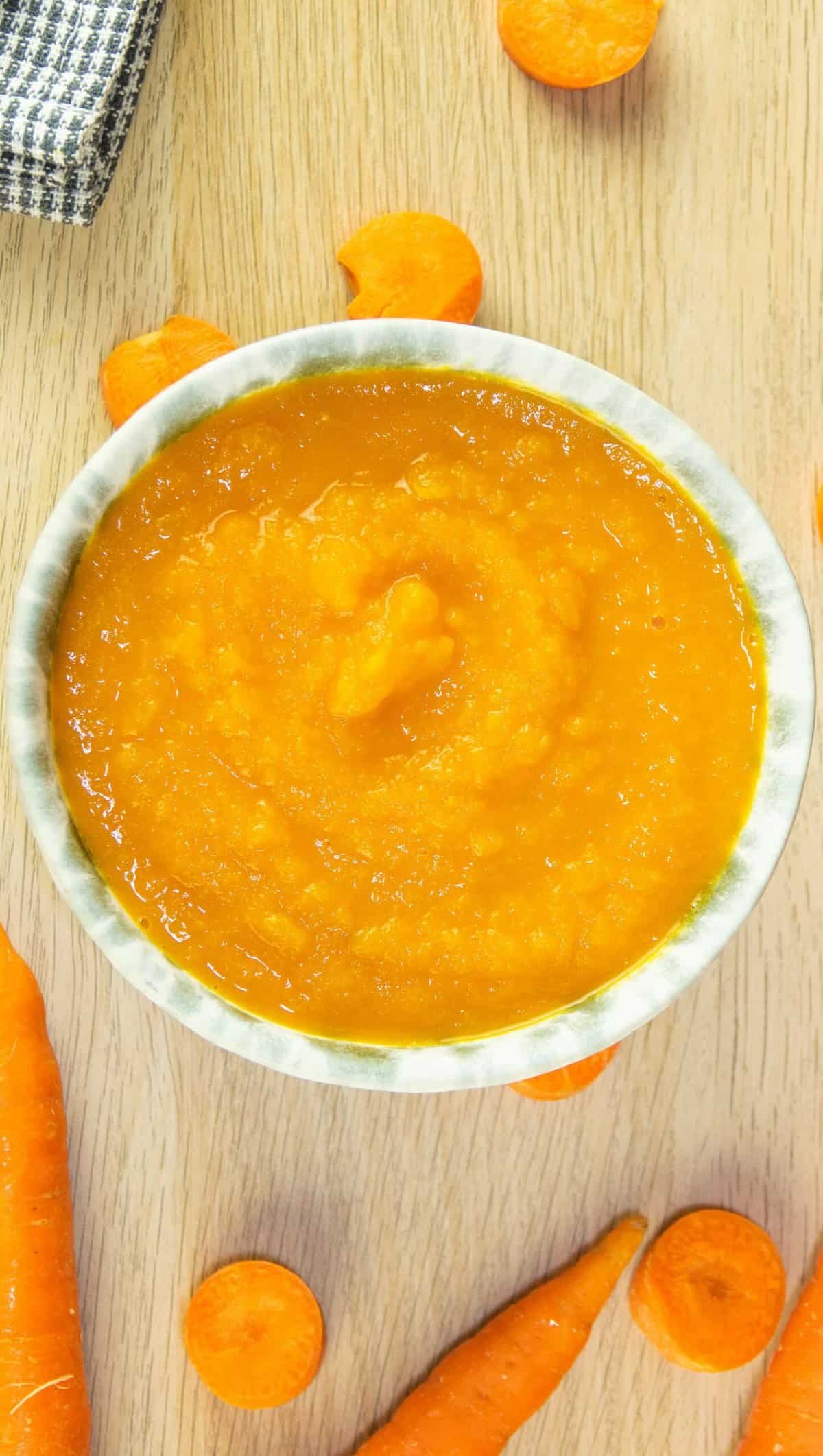 A bowl of carrot soup made with just 3 ingredients on a wooden board background with a few carrot pieces around the edges