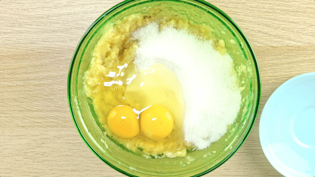 Sugar and eggs being added to mashed banana in a green-tinted glass bowl