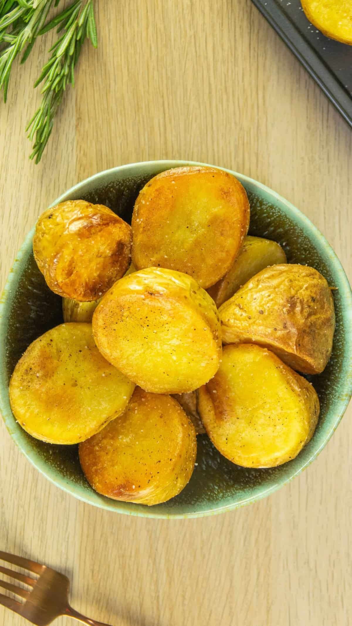 Bowl of roasted potato halves on a wooden background with a sprig of rosemary